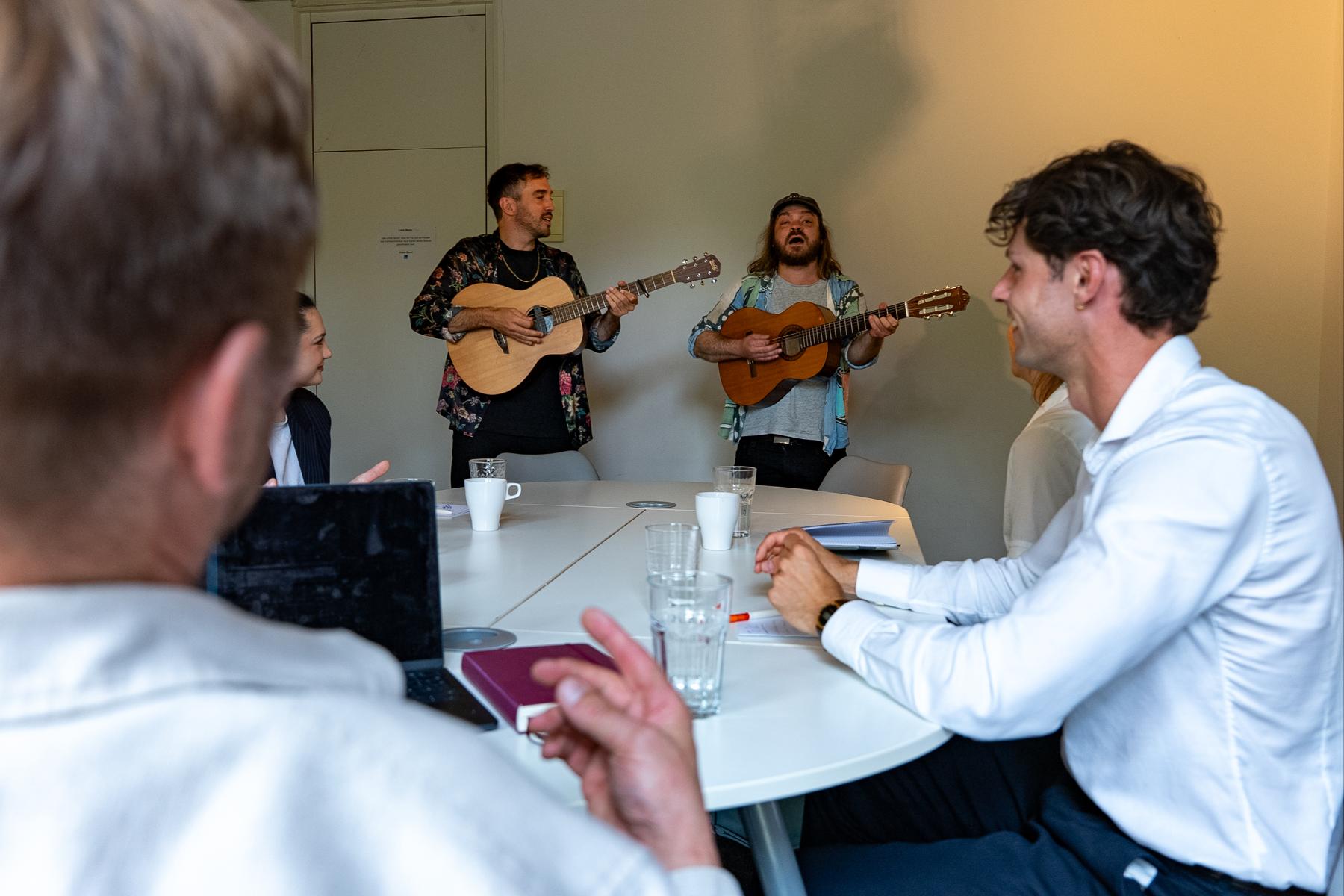 Zwei Musiker spielen Gitarre in einem Meetingraum, das Team am Tisch schaut zu